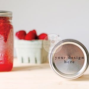 May include: A silver jar lid with the text "your design here" printed on it. The lid is sitting on a light wood surface. A jar of red jam and a bowl of strawberries are in the background.