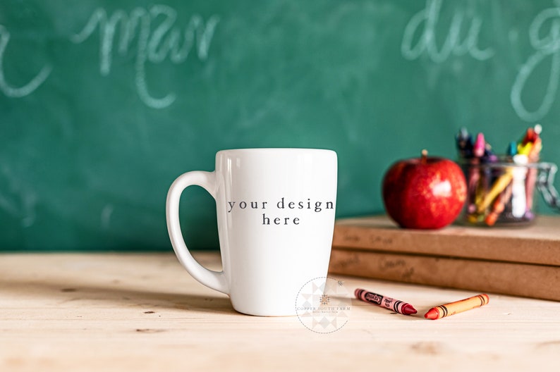 Op de afbeelding: Een witte keramische koffiemok met een zwarte inscriptie die "your design here" luidt. De mok staat op een houten tafel voor een groen schoolbord met witte krijtstreken. Op de achtergrond is een rode appel en een pot met kleurpotloden te zien.