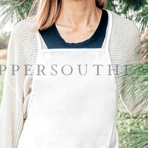 Op de afbeelding: Een witte schort met verstelbare banden, gedragen over een zwart shirt en een cr&egrave;mekleurige trui. De schort is leeg en kan worden gepersonaliseerd.