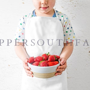 Pu&ograve; includere: Un bambino con un grembiule bianco e una camicia a pois colorati tiene una ciotola di fragole rosse fresche. Il testo "COPPERSOUTH FARM" &egrave; visibile sullo sfondo.