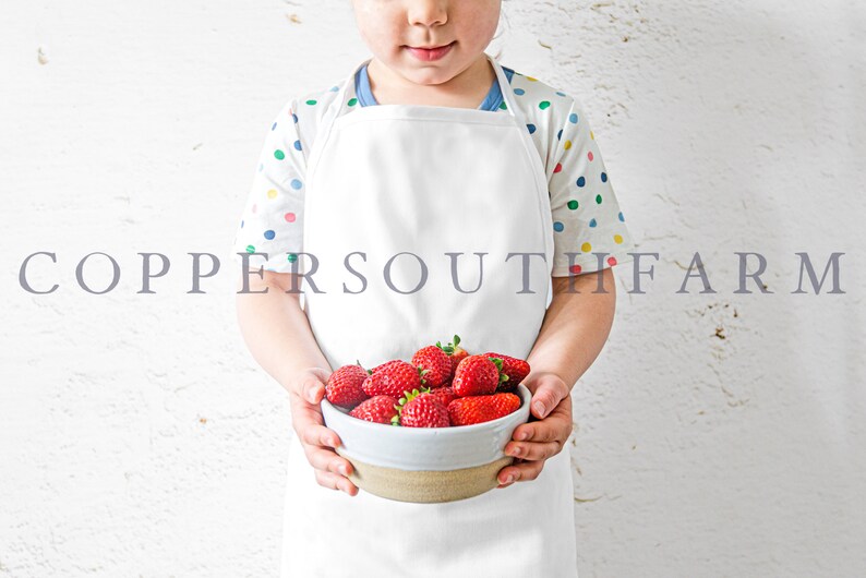 Pu&ograve; includere: Un bambino con un grembiule bianco e una camicia a pois colorati tiene una ciotola di fragole rosse fresche. Il testo "COPPERSOUTH FARM" &egrave; visibile sullo sfondo.
