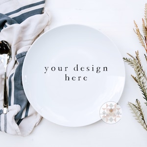 May include: A white ceramic plate with the text "your design here" on a white surface. A blue and white striped kitchen towel with a fork and spoon is to the left of the plate. A sprig of dried foliage is to the right of the plate.