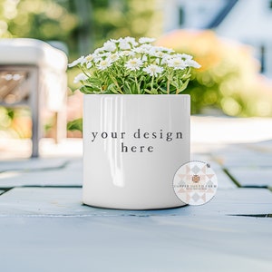 Op de afbeelding: Een witte keramische plantenpot met de tekst "your design here" aan de voorkant. De pot is gevuld met witte bloemen en staat op een grijs terras.