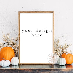 May include: A brown wooden frame with a white background and the text "your design here". The frame is sitting on a dark brown wooden surface with two orange pumpkins and two white pumpkins in front of it. There are dried flower stems on either side of the frame.