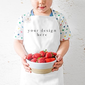Pu&ograve; includere: Un grembiule bianco con il testo "your design here" sul davanti. Un bambino indossa il grembiule e tiene una ciotola di fragole fresche.