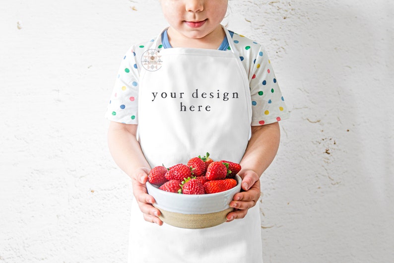 Pu&ograve; includere: Un grembiule bianco con il testo "your design here" sul davanti. Un bambino indossa il grembiule e tiene una ciotola di fragole fresche.