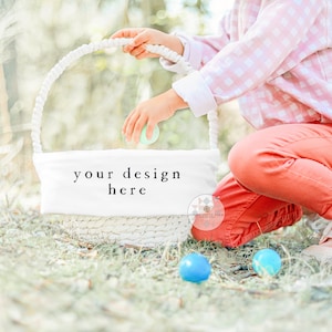 May include: A child wearing a pink and white gingham shirt and red trousers is kneeling in a grassy area. They are holding a white wicker basket with a white fabric liner that says "your design here". There are blue and green Easter eggs on the ground.