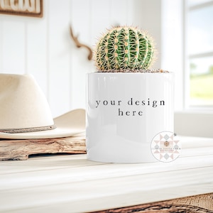 May include: A white ceramic planter with a green cactus, featuring the text "your design here." A beige cowboy hat rests on a piece of wood. The scene is set on a light-colored wooden table, with a window in the background.