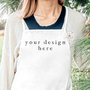 Op de afbeelding: Een witte schort met de tekst "your design here" erop gedrukt. De schort wordt gedragen door een persoon die voor een groene struik staat.