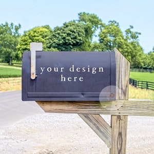 May include: A black mailbox with a wooden post, set against a backdrop of green fields and trees. The mailbox has the text "your design here" written on it.