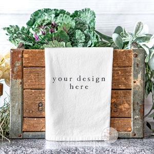 May include: White kitchen towel with the text "your design here" displayed in black lettering. The towel is folded and placed in a wooden crate with green plants on either side.