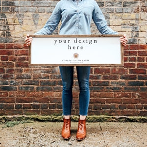 May include: A person holding a white sign with the text "your design here" and the words "Copper South Farm" in a brown wooden frame. The sign is in front of a brick wall.