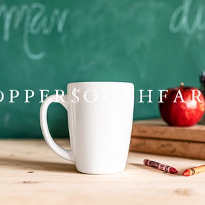 Op de afbeelding: Een witte keramische mok met een handvat, die op een houten tafel staat voor een groen schoolbord met witte krijtstreken. Op het schoolbord staan ook een rode appel en een pot met kleurpotloden.