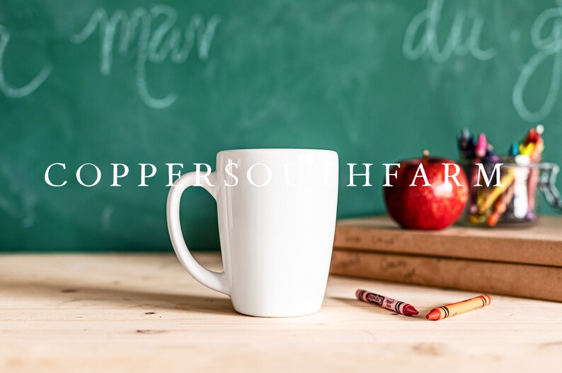 Op de afbeelding: Een witte keramische mok met een handvat, die op een houten tafel staat voor een groen schoolbord met witte krijtstreken. Op het schoolbord staan ook een rode appel en een pot met kleurpotloden.