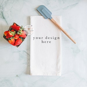 May include: A white kitchen towel with the text "your design here" on a marble surface. A blue silicone spatula with a wooden handle and a small wooden box filled with red strawberries are also on the surface.