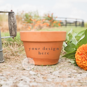 May include: A terracotta flower pot with the text "your design here" is displayed outdoors. An orange zinnia flower and a galvanized metal watering can are also in the image. The scene is set on a gravel path with a blurred background.