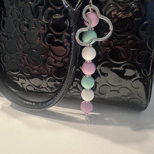 Puede incluir: Un llavero de cuentas con un dije de cabeza de Mickey Mouse plateado. El llavero presenta cuentas de colores pastel alternados, rosa, verde menta y blanco. El llavero est&aacute; sujeto a un bolso de mano de charol negro.