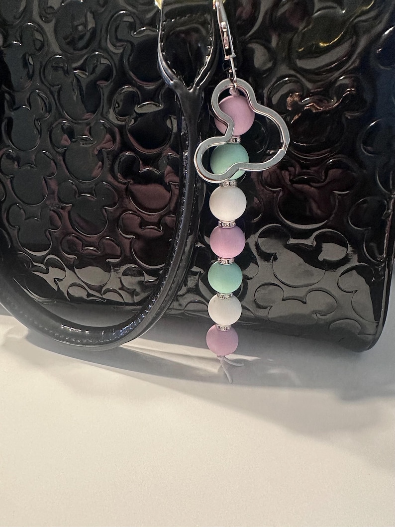 Puede incluir: Un llavero de cuentas con un dije de cabeza de Mickey Mouse plateado. El llavero presenta cuentas de colores pastel alternados, rosa, verde menta y blanco. El llavero est&aacute; sujeto a un bolso de mano de charol negro.