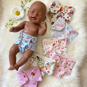 Könnte beinhalten: Eine Auswahl an bunten Puppenwindeln mit verschiedenen Mustern, darunter Regenbogen, Cowboys, Einhörner und Blumen. Die Stoffwindeln haben Druckknöpfe. Eine Puppe trägt eine der Windeln.