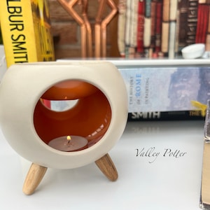 May include: A round, off-white ceramic wax warmer with a smooth matte exterior and a glossy orange interior. The vessel features a wide circular opening, a top dish for wax, and three light-coloured wooden legs. A small lit tea light candle sits inside.