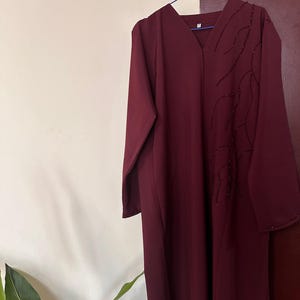 May include: A long, burgundy dress with a V-neck and long sleeves. The dress has a beaded embellishment on the front. It is displayed on a blue hanger. The background is a neutral colour.
