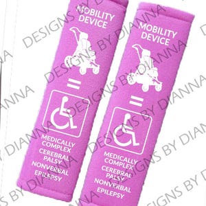 May include: Two pink mobility device straps with white text and graphics. Each strap features the words "MOBILITY DEVICE", a wheelchair symbol, and the phrases "MEDICALLY COMPLEX CEREBRAL PALSY NONVERBAL EPILEPSY".