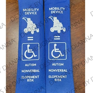 May include: Two blue fabric seatbelt covers with white text and symbols. The text reads "Mobility Device = Autism Nonverbal Elopement Risk". The symbol is a wheelchair.