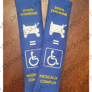 May include: Two blue fabric seatbelt covers with yellow text. The top cover reads "Down Syndrome" and the bottom cover reads "Medically Complex". Both covers feature a white wheelchair symbol and a white wagon symbol.