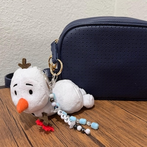 Op de afbeelding: Een marineblauwe geperforeerde schoudertas met een goudkleurig Mickey Mouse-embleem en een witte pluche sneeuwpop-sleutelhanger aan de riem. De sleutelhanger heeft een oranje neus en een kralensnoer met de letters die de naam OLAF vormen.