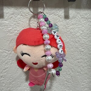 Op de afbeelding: Een pluche sleutelhanger met een figuurtje met felrood haar en een roze jurk, bevestigd aan een zilveren ring. Aan de ring hangt een kralensnoer met witte, roze, paarse en groene kralen, letterkralen die ARIEL spellen en een kleine zilveren vorkbedel.