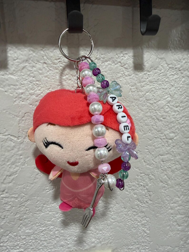 Op de afbeelding: Een pluche sleutelhanger met een figuurtje met felrood haar en een roze jurk, bevestigd aan een zilveren ring. Aan de ring hangt een kralensnoer met witte, roze, paarse en groene kralen, letterkralen die ARIEL spellen en een kleine zilveren vorkbedel.
