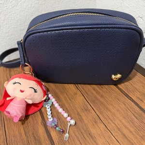 Op de afbeelding: Een marineblauwe geperforeerde schoudertas met een gouden rits en een klein gouden embleem. Aan de tas hangt een pluche sleutelhanger van een figuurtje met rood haar en een roze jurk, met een kralenketting met de naam ARIEL en een vorkbedeltje.
