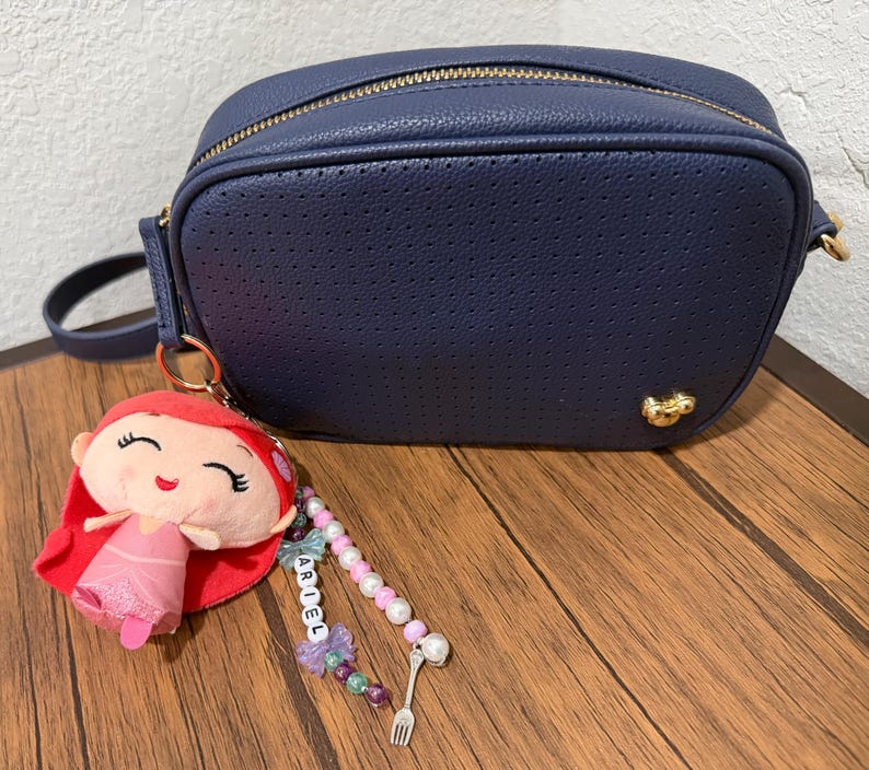Op de afbeelding: Een marineblauwe geperforeerde schoudertas met een gouden rits en een klein gouden embleem. Aan de tas hangt een pluche sleutelhanger van een figuurtje met rood haar en een roze jurk, met een kralenketting met de naam ARIEL en een vorkbedeltje.