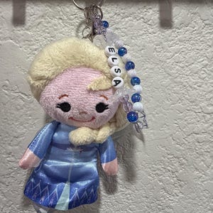 Peut inclure: Un porte-cl&eacute;s en peluche repr&eacute;sentant un personnage aux cheveux blonds v&ecirc;tu d'une robe bleue, fix&eacute; &agrave; un anneau m&eacute;tallique. Un cordon de perles est suspendu &agrave; l'anneau, avec des perles blanches formant le nom ELSA, altern&eacute;es de perles bleues et transparentes.