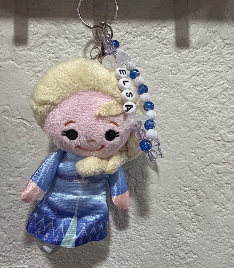 Peut inclure: Un porte-cl&eacute;s en peluche repr&eacute;sentant un personnage aux cheveux blonds v&ecirc;tu d'une robe bleue, fix&eacute; &agrave; un anneau m&eacute;tallique. Un cordon de perles est suspendu &agrave; l'anneau, avec des perles blanches formant le nom ELSA, altern&eacute;es de perles bleues et transparentes.