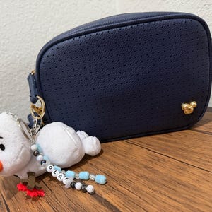 Op de afbeelding: Een marineblauwe geperforeerde schoudertas met een goudkleurig Mickey Mouse-embleem. Aan de zijring hangt een pluche sneeuwpop-sleutelhanger met een oranje neus, bruine takarmpjes en een kralensnoer met de letters die de naam OLAF vormen.