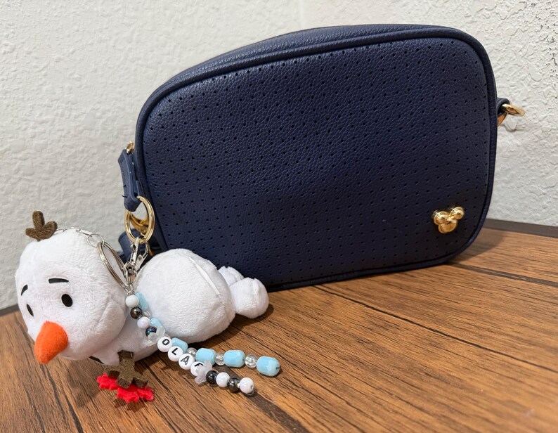 Op de afbeelding: Een marineblauwe geperforeerde schoudertas met een goudkleurig Mickey Mouse-embleem. Aan de zijring hangt een pluche sneeuwpop-sleutelhanger met een oranje neus, bruine takarmpjes en een kralensnoer met de letters die de naam OLAF vormen.