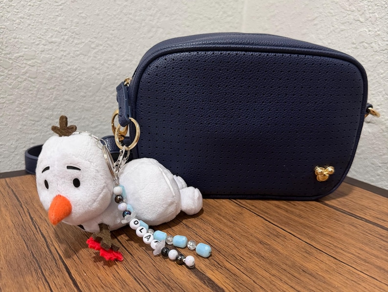 Op de afbeelding: Een marineblauwe geperforeerde schoudertas met een goudkleurig Mickey Mouse-embleem en een witte pluche sneeuwpop-sleutelhanger aan de riem. De sleutelhanger heeft een oranje neus en een kralensnoer met de letters die de naam OLAF vormen.