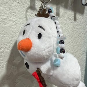 Op de afbeelding: Een pluche sneeuwpop-sleutelhanger met een wit lichaam, een feloranje viltneus, zwarte viltwenkbrauwen en een bruine vilt-takarm. Een zilveren metalen ring aan de bovenkant is versierd met een streng witte, lichtblauwe en donkere kralen.
