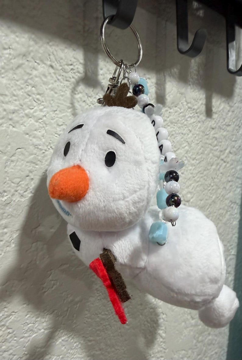 Op de afbeelding: Een pluche sneeuwpop-sleutelhanger met een wit lichaam, een feloranje viltneus, zwarte viltwenkbrauwen en een bruine vilt-takarm. Een zilveren metalen ring aan de bovenkant is versierd met een streng witte, lichtblauwe en donkere kralen.