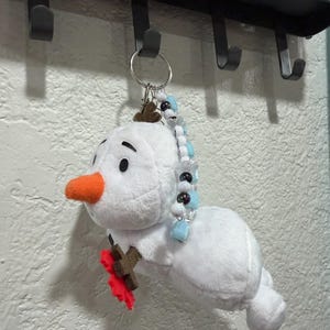 Op de afbeelding: Een pluche sneeuwpop-sleutelhanger met een wit lichaam, een feloranje viltneus en bruine vilten takarmen. E&eacute;n arm houdt een kleine rode viltbloem vast. Aan de bovenkant zit een zilveren metalen sleutelring versierd met blauwe, witte en zwarte kralen.