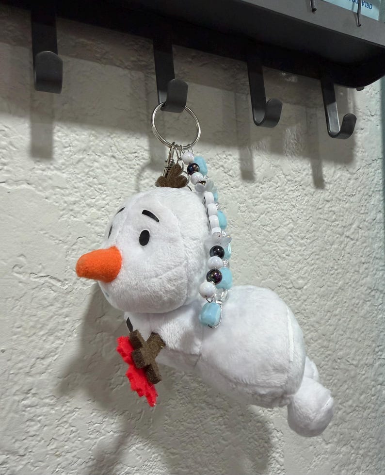 Op de afbeelding: Een pluche sneeuwpop-sleutelhanger met een wit lichaam, een feloranje viltneus en bruine vilten takarmen. E&eacute;n arm houdt een kleine rode viltbloem vast. Aan de bovenkant zit een zilveren metalen sleutelring versierd met blauwe, witte en zwarte kralen.