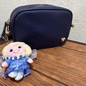 Peut inclure: Un sac bandouli&egrave;re rectangulaire bleu marine avec une texture perfor&eacute;e et une petite ic&ocirc;ne Mickey Mouse dor&eacute;e dans le coin inf&eacute;rieur droit. Un porte-cl&eacute;s poup&eacute;e en peluche repr&eacute;sentant un personnage blond en robe bleue est attach&eacute; au sac.