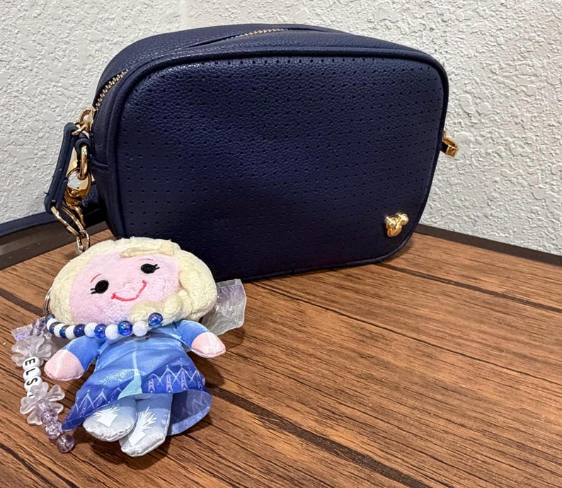 Peut inclure: Un sac bandouli&egrave;re rectangulaire bleu marine avec une texture perfor&eacute;e et une petite ic&ocirc;ne Mickey Mouse dor&eacute;e dans le coin inf&eacute;rieur droit. Un porte-cl&eacute;s poup&eacute;e en peluche repr&eacute;sentant un personnage blond en robe bleue est attach&eacute; au sac.