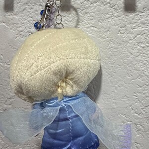Peut inclure: Un porte-cl&eacute;s en peluche repr&eacute;sentant un personnage aux cheveux blonds tress&eacute;s, v&ecirc;tu d'une robe en satin bleu avec une cape bleu clair transparente. La figurine est fix&eacute;e &agrave; un anneau en m&eacute;tal argent&eacute; avec une petite cha&icirc;ne de perles bleues et blanches.