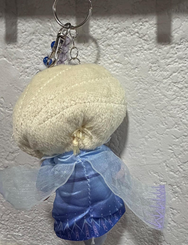 Peut inclure: Un porte-cl&eacute;s en peluche repr&eacute;sentant un personnage aux cheveux blonds tress&eacute;s, v&ecirc;tu d'une robe en satin bleu avec une cape bleu clair transparente. La figurine est fix&eacute;e &agrave; un anneau en m&eacute;tal argent&eacute; avec une petite cha&icirc;ne de perles bleues et blanches.