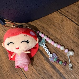 Op de afbeelding: Een sleutelhanger met een pluche pop met rood haar en een roze jurk, bevestigd aan een kralensnoer met witte en paarse kralen, een bedel in de vorm van een vork en witte letterkralen die verticaal de naam ARIEL spellen.