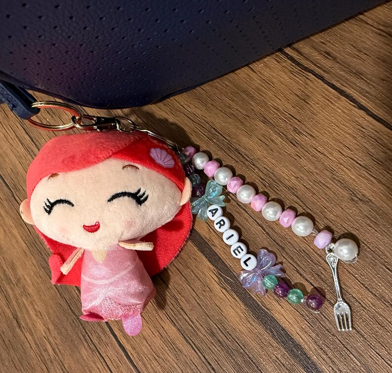 Op de afbeelding: Een sleutelhanger met een pluche pop met rood haar en een roze jurk, bevestigd aan een kralensnoer met witte en paarse kralen, een bedel in de vorm van een vork en witte letterkralen die verticaal de naam ARIEL spellen.