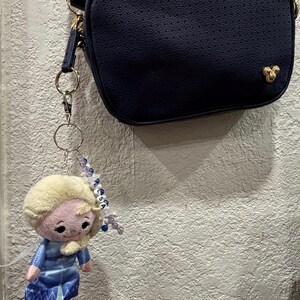 Peut inclure: Un sac bandouli&egrave;re bleu marine avec une petite ic&ocirc;ne dor&eacute;e Mickey Mouse attach&eacute; &agrave; un porte-cl&eacute;s en peluche repr&eacute;sentant Elsa. Le porte-cl&eacute;s comprend un anneau en m&eacute;tal argent&eacute;, des perles bleues et blanches et des lettres formant ELSA.