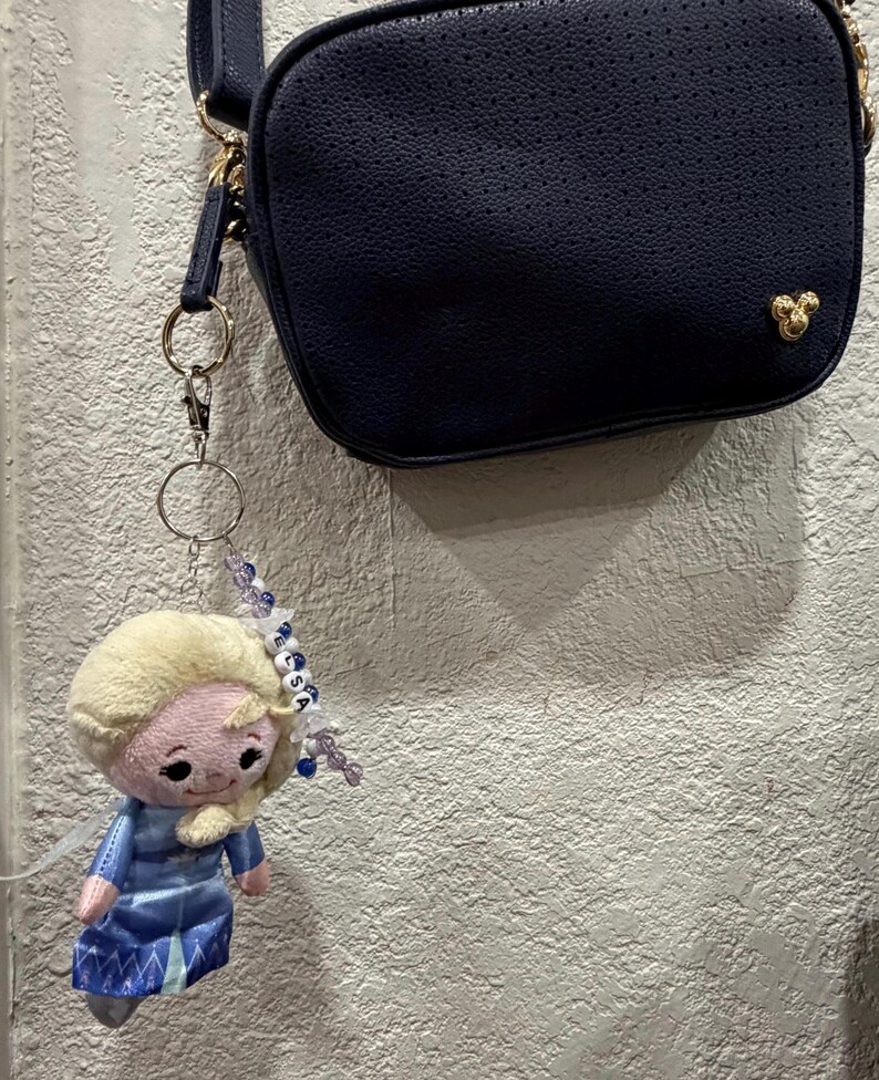 Peut inclure: Un sac bandouli&egrave;re bleu marine avec une petite ic&ocirc;ne dor&eacute;e Mickey Mouse attach&eacute; &agrave; un porte-cl&eacute;s en peluche repr&eacute;sentant Elsa. Le porte-cl&eacute;s comprend un anneau en m&eacute;tal argent&eacute;, des perles bleues et blanches et des lettres formant ELSA.